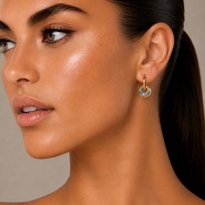 May include: A close-up of a person wearing a gold hoop earring with a circular, blue-grey stone inlay. The earring is a small, delicate piece of jewellery. The person has dark hair pulled back in a ponytail and is looking to the left.