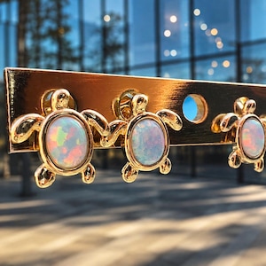 May include: Three gold-toned turtle-shaped earrings attached to a gold display card. Each turtle features a central oval-shaped iridescent opal cabochon with flashes of pink, orange, and blue. The metal has a polished finish and detailed flippers.