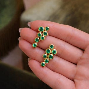May include: A pair of gold-colored stud earrings, each featuring a cluster of vibrant green gemstones. The gemstones are round and set in a bezel setting. The earrings are displayed on a light-toned background.