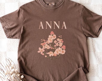 May include: A brown t-shirt with the name "ANNA" printed above a floral design. The floral design features pink and peach-coloured flowers. The shirt is displayed on a white checkered surface with a bundle of dried flowers.