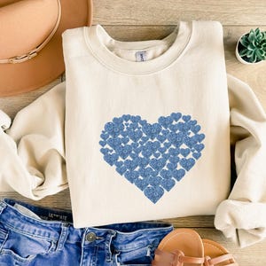 May include: Cream-colored sweatshirt featuring a large heart design made of smaller blue hearts. The sweatshirt is paired with blue jeans, a tan hat, and brown sandals. A small succulent plant in a white pot is also visible.