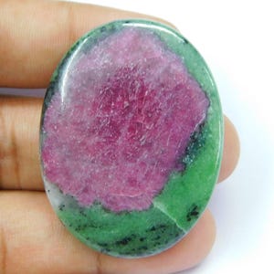 Op de afbeelding: Een ovaalvormige cabochon edelsteen met een levendig magenta-roze centrum omringd door een gevlekte groene en zwarte mineraalmatrix. De steen heeft een glad, gepolijst oppervlak dat het natuurlijke kleurcontrast tussen de mineralen benadrukt.