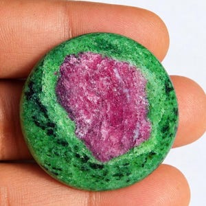 May include: A circular polished gemstone featuring a vibrant magenta-pink center surrounded by a textured green and black zoisite matrix. The stone has a smooth, rounded surface with a distinct contrast between the crystalline core and the outer rim.