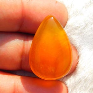 May include: A polished, teardrop-shaped gemstone in shades of orange and yellow. The translucent stone has a smooth, glossy surface and is held in a person's hand against a white, fluffy background. The gemstone is likely for jewelry making.