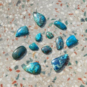 May include: A selection of polished gemstones in teal, turquoise, and deep blue hues. The stones, in various shapes from oval to teardrop, are arranged on a speckled grey surface. The gemstones have a marbled appearance with darker blue and green inclusions.