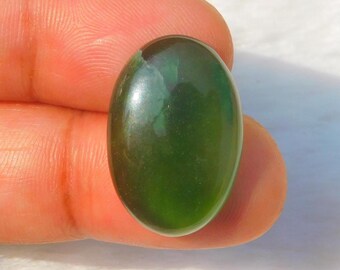 May include: A polished, oval-shaped, dark green gemstone. The stone has a smooth, reflective surface and is held between two fingers. The gemstone's color varies from a deep, forest green to a lighter, translucent green.