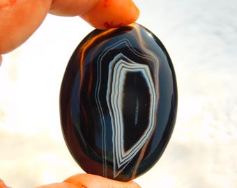 May include: A polished, oval-shaped agate stone. The stone features a striking pattern of concentric bands in shades of black, brown, white, and orange. The agate is held between fingers, showcasing its smooth, glossy surface and natural beauty.