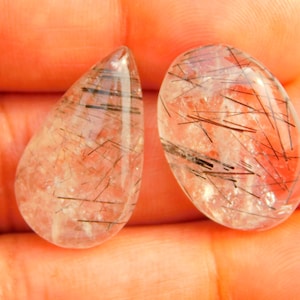 May include: Two clear, polished gemstones with internal black inclusions. One is teardrop-shaped, the other oval. The stones have a translucent quality, with reddish and black linear patterns visible.