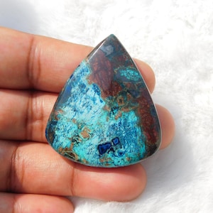 May include: A polished, triangular gemstone with a mix of blue, teal, and brown hues. The stone has a smooth, glossy surface and a unique pattern of colors and textures. The gemstone is held in a hand.