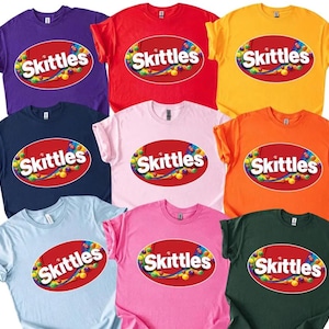May include: A collection of colorful t-shirts in various colors, including purple, red, yellow, navy, pink, orange, light blue, and green. Each shirt features the Skittles logo in a red oval with colorful candies.