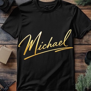 May include: Black t-shirt featuring the name "Michael" in a gold, cursive script. The text is the focal point of the design, set against the dark fabric. The shirt has short sleeves and a classic crew neck.