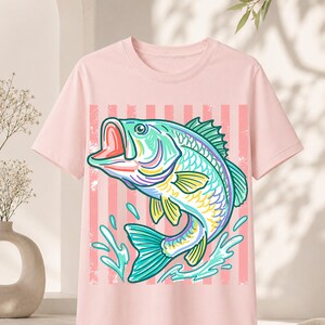 May include: A light pink t-shirt featuring a colorful illustration of a fish jumping out of the water. The fish is primarily teal, yellow, and purple, with water splashes and a pink striped background.