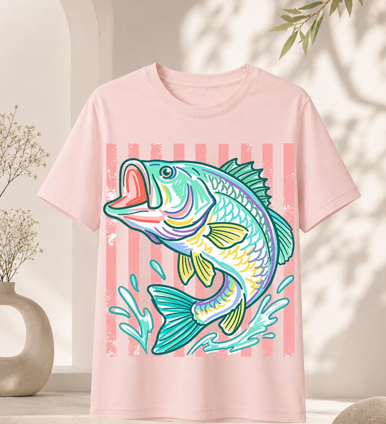 May include: A light pink t-shirt featuring a colorful illustration of a fish jumping out of the water. The fish is primarily teal, yellow, and purple, with water splashes and a pink striped background.