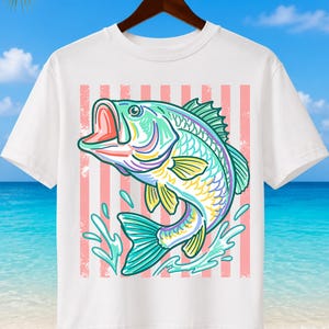 May include: White t-shirt featuring a colorful illustration of a largemouth bass jumping out of water. The fish is depicted in shades of teal, yellow, purple, and pink, set against a background of pink and white vertical stripes. The shirt is hanging on a wooden hanger.