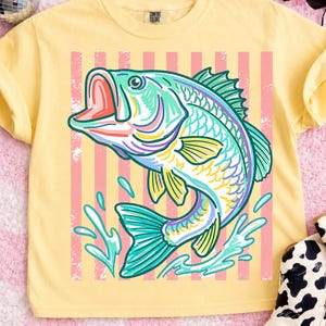 May include: Yellow Comfort Colors t-shirt featuring a colorful illustration of a leaping fish. The fish is depicted in shades of teal, purple, yellow, and pink, set against a background of pink stripes. The shirt has a classic crew neck.