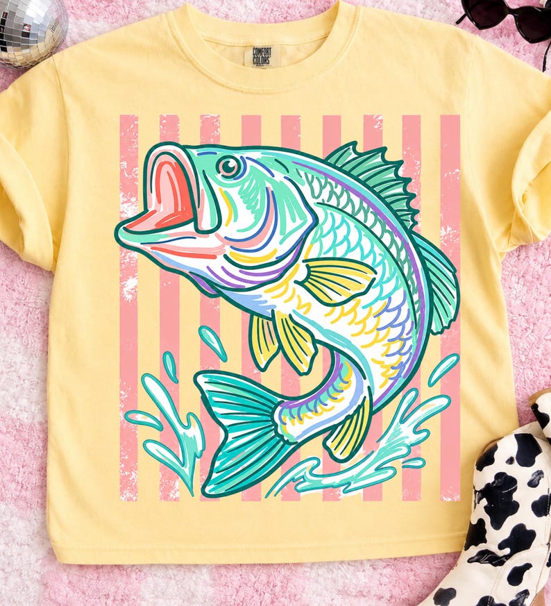 May include: Yellow Comfort Colors t-shirt featuring a colorful illustration of a leaping fish. The fish is depicted in shades of teal, purple, yellow, and pink, set against a background of pink stripes. The shirt has a classic crew neck.