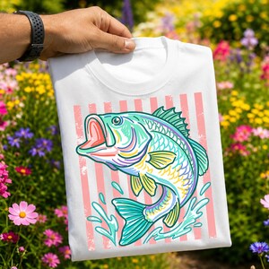 May include: White t-shirt featuring a colorful illustration of a jumping fish with an open mouth. The fish is in shades of teal, yellow, purple, and pink, set against a background of pink vertical stripes. The shirt is held up by a person.