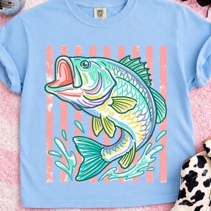 May include: Light blue t-shirt featuring a colorful illustration of a largemouth bass jumping out of water. The fish is depicted with vibrant hues of green, yellow, purple, and pink. Vertical pink stripes are behind the fish. The shirt has a classic crew neck.