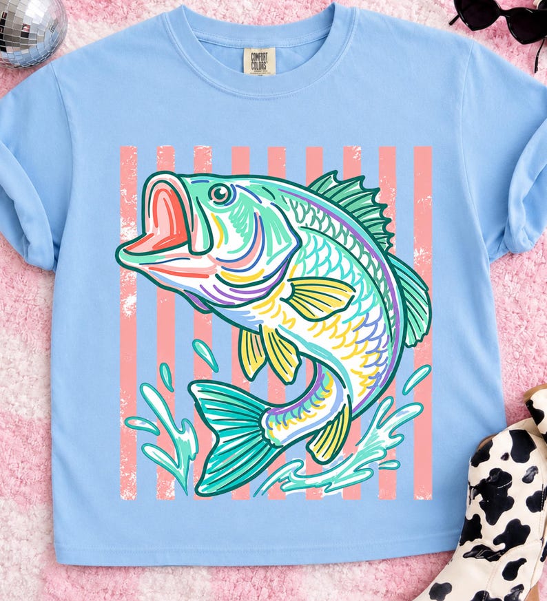 May include: Light blue t-shirt featuring a colorful illustration of a largemouth bass jumping out of water. The fish is depicted with vibrant hues of green, yellow, purple, and pink. Vertical pink stripes are behind the fish. The shirt has a classic crew neck.