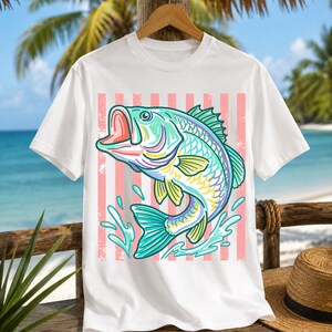May include: White t-shirt featuring a colorful illustration of a fish with an open mouth, splashing water, and a pink striped background. The fish has teal, yellow, purple, and green details. The shirt is hanging on a wooden hanger.