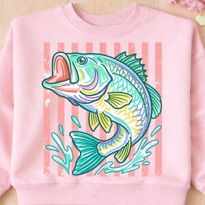 May include: A light pink sweatshirt featuring a colorful illustration of a leaping fish. The fish is depicted in shades of teal, yellow, purple, and green, with water splashes and a striped background. The design has a vintage, hand-drawn aesthetic.