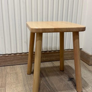 Op de afbeelding: Een kleine, lichtgekleurde houten kruk met een vierkant blad met afgeronde hoeken en vier stevige, cilindrische poten. De kruk is gemaakt van glad, natuurlijk gekleurd hout met een zichtbare houtnerf en staat op een vlakke ondergrond.