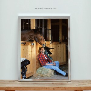 May include: A framed image of a horse in a stable with a person sitting on a hay bale. The horse is brown, and the person is wearing a red and black plaid shirt and blue jeans. The image is on a wooden table.