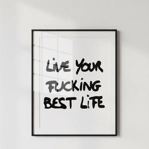 以下が含まれることがあります： 黒いフレームのアートプリント。手書き風の黒い文字で「Live Your Fucking Best Life」と書かれています。白い背景にテキストが配置されています。白い壁に飾られたアートワークです。