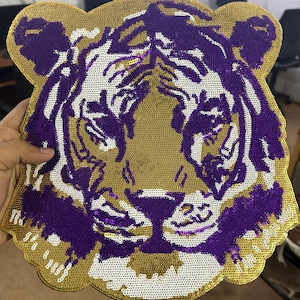 Op de afbeelding: Een grote, met pailletten bezette tijgerkop-patch in goud, paars en wit. Het gezicht van de tijger is gedetailleerd met paarse strepen en witte accenten, tegen een gouden achtergrond. De patch wordt door een hand vastgehouden.