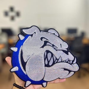 Peut inclure: Un grand écusson représentant une tête de bouledogue. Le bouledogue est principalement argenté avec une bordure bleue et des détails noirs. L'écusson est tenu dans une main, mettant en valeur sa taille et ses détails. Idéal pour les vêtements ou accessoires.