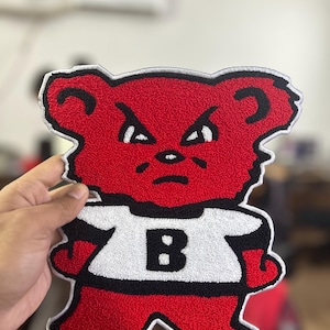 Peut inclure: Écusson en forme d'ours rouge avec un t-shirt blanc portant un "B" noir. L'ours a une expression fâchée et un contour noir. L'écusson est destiné aux vêtements ou accessoires.