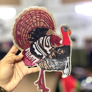 May include: A detailed turkey patch with a decorative design. The patch features a fan of brown and red feathers, a black and white body, and red legs. The edges are outlined in gold. The patch is held up by a hand.
