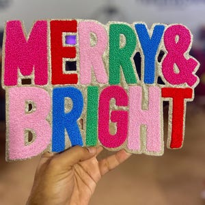 May include: A colorful sign with the words "MERRY & BRIGHT" in pink, red, green, and blue, with a gold glitter border. The sign is held by a hand, and is a festive decoration.
