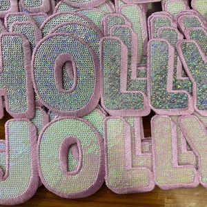 Può includere: Una collezione di toppe ricamate che compongono la scritta "HOLLY JOLLY" in maiuscolo. Le lettere hanno un riempimento di paillettes argentate e sono bordate di rosa. Le toppe sono sparse su una superficie.