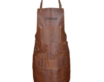 May include: A brown leather apron with multiple pockets and adjustable straps. The apron has the text "DEMHAAS" printed on the chest area. It features a loop for hanging tools and is designed for use in the kitchen or workshop.