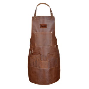 Peut inclure: Tablier en cuir marron avec plusieurs poches et une boucle. Le tablier a une sangle de cou et une fermeture à boucle. Le mot "NAME" est imprimé sur un petit patch rectangulaire.