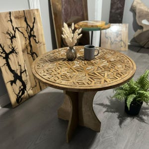 May include: A round, light brown wooden table with a carved geometric pattern on the tabletop. The table has a pedestal base and is surrounded by other wooden art pieces. A gray mug and a small vase with dried flowers sit on the table.