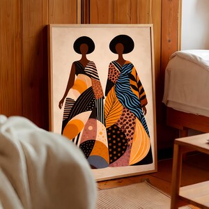 May include: A framed art print featuring two stylized figures in colorful, patterned dresses. The artwork showcases a palette of orange, black, blue, and brown, with geometric and organic shapes. The figures have dark skin and wear large, black afro hairstyles.