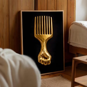 May include: A framed artwork featuring a gold-colored Afro pick held by a clenched fist against a black background. The artwork is displayed in a room with wooden paneling and a bed.