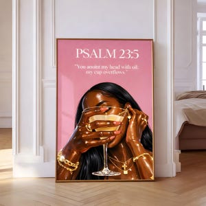 May include: A framed art print with a pink background, featuring a woman holding a cocktail glass. The artwork includes the text "PSALM 23:5" and a quote. The woman is wearing gold jewellery, including bracelets, rings, and a necklace.