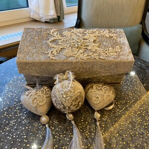 Könnte beinhalten: Dekorative Box und Ornamente in neutralen Farben. Die rechteckige Box ist mit einem strukturierten Stoff bezogen und mit aufwendiger goldener Spitze verziert. Drei Ornamente, darunter Herz- und Kugelformen, weisen ähnliche Verzierungen und silberne Quasten auf.