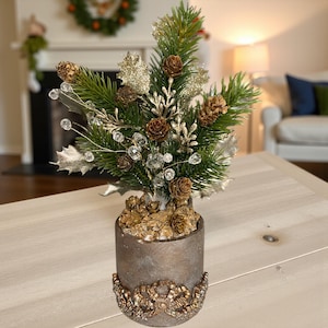 May include: A decorative Christmas tree arrangement in a cylindrical pot. The tree features green pine needles, brown pine cones, silver leaves, and sparkling crystal accents. The pot is a muted bronze color with a gold decorative base.