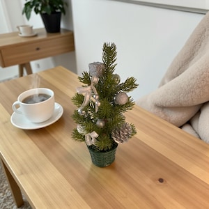 Könnte beinhalten: Ein kleiner, künstlicher Weihnachtsbaum in einem grünen Topf, geschmückt mit silbernen Ornamenten, Schleifen und einem Tannenzapfen. Eine Tasse Kaffee steht auf einer Untertasse auf einem Holztisch.