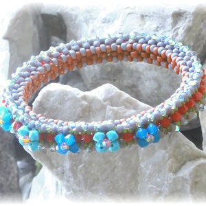 May include: A beaded bracelet with a multi-coloured design. The bracelet is made with small, round beads in shades of purple, orange, and red. The beads are arranged in a pattern that creates a flower design. The bracelet is accented with blue beads that create a flower design.
