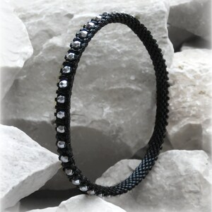 May include: A black beaded bracelet with silver-colored accents. The bracelet is made of small, dark beads and features a row of faceted silver beads. The bracelet is displayed against a white, textured background.