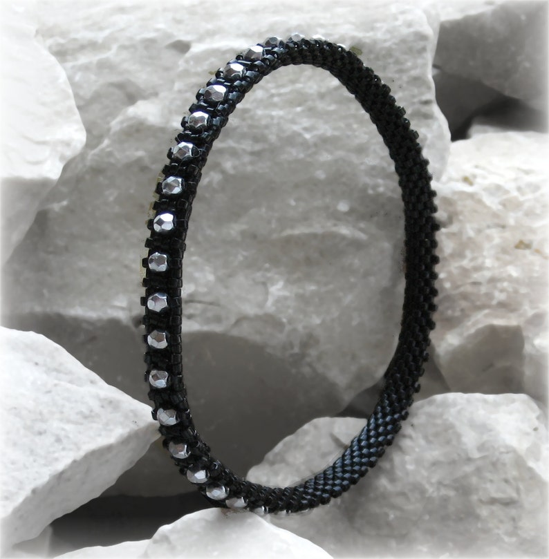 May include: A black beaded bracelet with silver-colored accents. The bracelet is made of small, dark beads and features a row of faceted silver beads. The bracelet is displayed against a white, textured background.