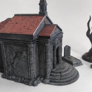 Può includere: Un modello in miniatura di una cappella in pietra con tetto di tegole rosse. La cappella presenta dettagli architettonici, tra cui colonne, un piccolo campanile e intagli decorativi. Il modello è dipinto in tonalità di grigio e marrone, con un aspetto invecchiato. Sono visibili anche due lapidi in miniatura.