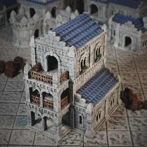 Pode incluir: Um edifício de pedra em miniatura com um telhado de telhas azuis e uma varanda com uma grade marrom. O edifício tem janelas arqueadas e faz parte de um conjunto de estruturas semelhantes, provavelmente para jogos de mesa ou modelagem.