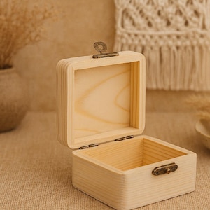 Puede incluir: Una caja de madera abierta, de color claro, con tapa con bisagras y un pequeño cierre metálico. La caja es cuadrada y tiene un acabado de veta de madera natural. El fondo incluye un jarrón con flores secas y un tapiz decorativo.