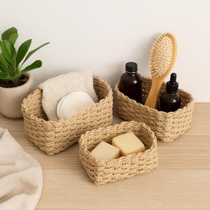 Puede incluir: Tres cestas rectangulares beige tejidas de varios tamaños. Una cesta contiene una tela doblada y discos de algodón redondos. Otra contiene un cepillo de pelo de madera y dos botellas marrones. La cesta más pequeña tiene dos pastillas de jabón. Una pequeña planta en maceta está en el fondo.